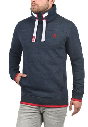 Sweatshirt bleu marine avec un col montant, des cordons de serrage blancs, des accents rouges et un logo sur la poitrine. Il présente une texture douce et des poches latérales.