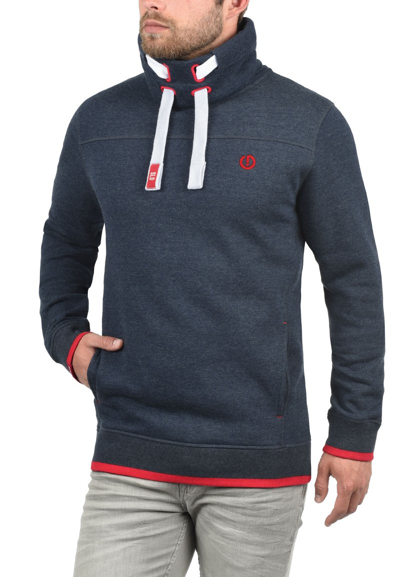 Sudadera azul marino con cuello alto, cordones blancos, acentos rojos y un logo en el pecho. Presenta una textura suave y bolsillos laterales.