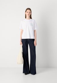 T-shirt en coton blanc avec un logo pastel sur la poitrine, associé à un jean en denim bleu foncé à jambes larges et à un sac fourre-tout beige.