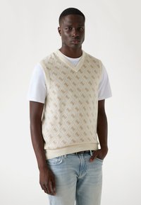 Strick-Weste in Creme mit einem Rautenmuster in Beige, ausgestattet mit einem V-Ausschnitt und einem gerippten Saum. Kombiniert mit einem weißen T-Shirt und hellblauen Jeans.