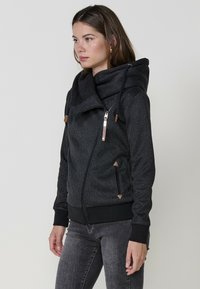 Felpa con zip nera, caratterizzata da un motivo testurizzato, un ampio colletto e cordini. Presenta una zip metallica e accenti marroni a contrasto.