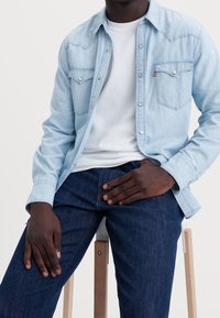 Jasnoniebieska koszula dżinsowa na zatrzaski, z dwoma kieszeniami na piersi, zestawiona z białym t-shirtem i ciemnoniebieskimi dżinsami, siedząca na stołku.