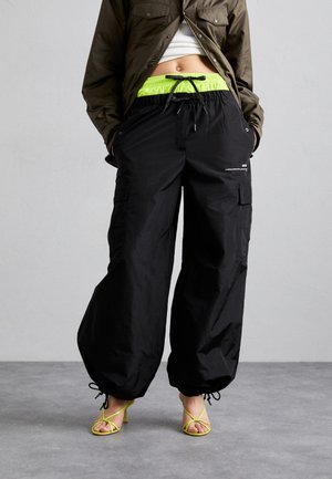 Person wearing loose black cargo pants with neon green drawstring waist, cropped olive jacket, and neon green strappy heels, standing on gray floor.