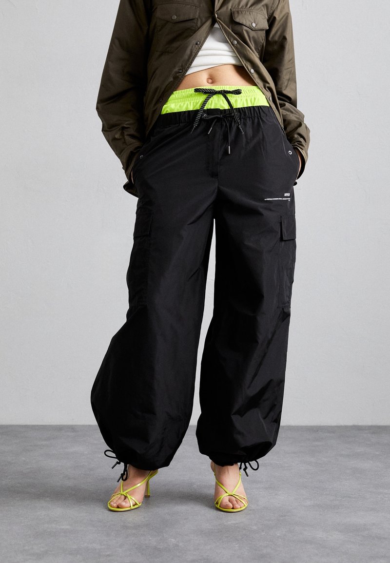Personne portant un pantalon cargo noir ample avec un cordon de serrage vert fluo à la taille, une veste olive courte et des talons à brides vert fluo, debout sur un sol gris.