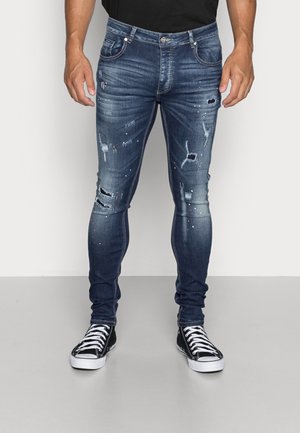 Mann trägt dunkelblaue Slim-Fit-Jeans mit Used-Look und Farbspritzern sowie schwarze High-Top-Sneaker, steht vor weißem Hintergrund.