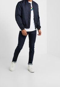 Námořnicky modrá bomber bunda s žebrovanými manžetami, nošená přes bílé tričko. Outfit doplňují tmavé džíny a bílé tenisky. Jednoduchý, čistý design.