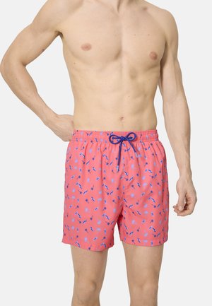 Hombre con pantalones cortos de baño color coral con pequeño patrón de palmeras azules y tablas de surf, y cordón azul marino, de pie contra un fondo liso.