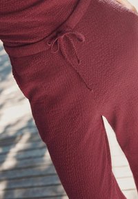 Maroonfarbene, strukturierte Sweatpants mit einem verstellbaren Kordelzugbund und weichem, gewebtem Stoff, seitlich auf einer Holzoberfläche dargestellt.