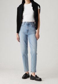 Jeans de tiro alto en un azul claro con detalles desgastados, combinados con una camiseta blanca corta y un cárdigan negro de canalé, terminando con unos mocasines negros.