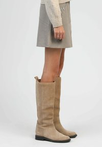Bottes en suede beige montantes avec un bout rond, un large tige et une semelle en caoutchouc noir. Texture lisse avec un minimum de détails de couture.