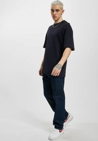 Marine oversized t-skjorte med korte ermer, kombinert med mørkeblå jeans og hvite joggesko med røde detaljer. Tatovering synlig på hånden.