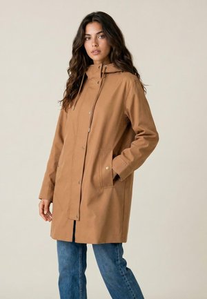 Jeune femme aux longs cheveux foncés portant un manteau à capuche beige avec des boutons-pression et un jean bleu, debout devant un fond clair uni.