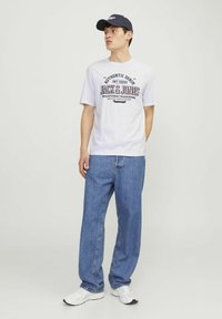 Hvid T-shirt med trykt logo og tekst, parret med lyseblå baggy denimjeans og hvide sneakers. Model iført en marineblå kasket.