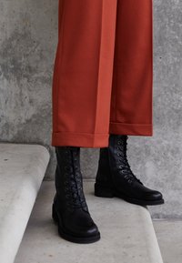 Bottes cheville en cuir noir à lacets avec un bout rond, associées à un pantalon ample de couleur rouille, mettant en valeur un ourlet retroussé.