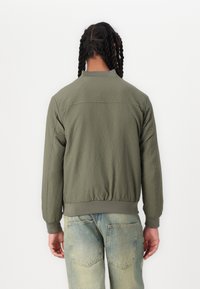 Zelena bomber jakna sa teksturom i uskim pojasom i rukavima, koja ima okrugli ovratnik i šivane detalje na leđima.