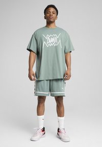Mintgrünes Oversized-T-Shirt mit weißem Grafikdruck, kombiniert mit passenden Netzshorts mit schwarzen Akzenten und PUMA-Logo. Weiße Socken und Sneakers.