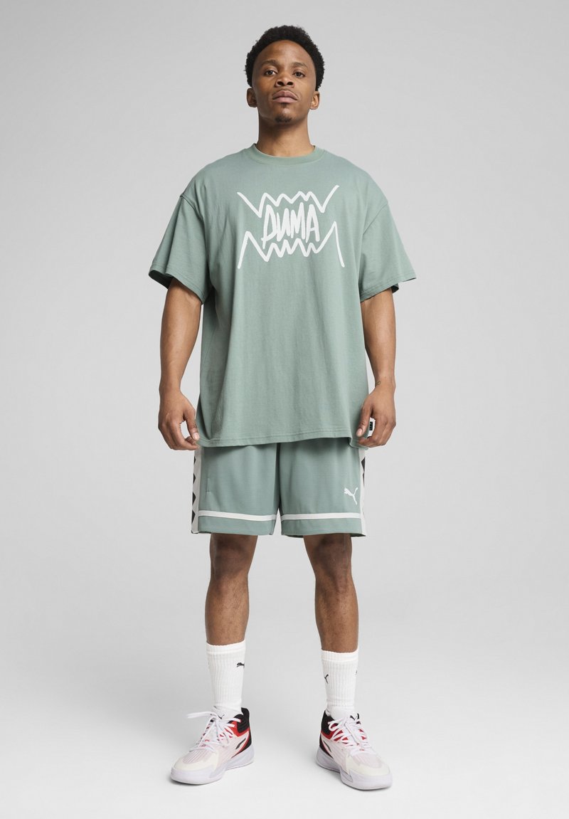 Mintgrünes Oversized-T-Shirt mit weißem Grafikdruck, kombiniert mit passenden Netzshorts mit schwarzen Akzenten und PUMA-Logo. Weiße Socken und Sneakers.