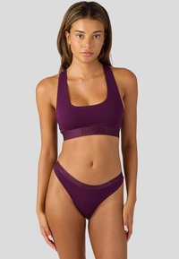 Kobieta ubrana w dopasowany fioletowy stanik sportowy i bieliznę z wysokim wycięciem, stojąca na jednolitym tle.