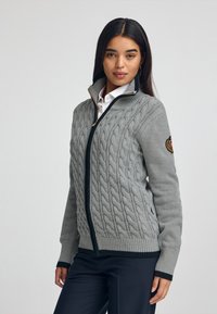 Grå kabelstickad zip-up tröja med en mörk dragkedja och ribbade muddarna, med ett cirkulärt märke på vänster ärm. Slät textur.
