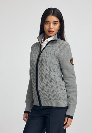 Grau gestrickter Zip-Up-Pullover mit dunklem Reißverschluss und gerippten Bündchen, versehen mit einem runden Aufnäher auf dem linken Ärmel. Glatte Textur.