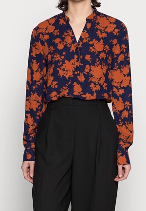 Blouse bleu marine à motif floral orange, manches longues et col en V, portée rentrée dans un pantalon noir taille haute et cintré.