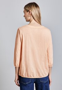Top ligero color durazno con cuello redondo y mangas tres cuartos. Presenta un dobladillo fruncido y una tela suave y texturizada.