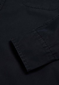 Camicia nera in cotone con maniche e tasca, caratterizzata da una texture liscia e una finitura pulita, che mette in evidenza cuciture doppie e bordi netti.