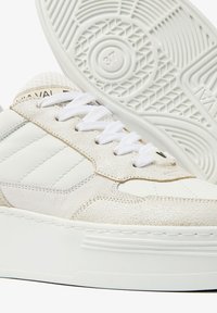 White sneakers featuring a mix of smooth leather and textured suede, with a rubber sole, flat laces, and a subtle logo on the heel.