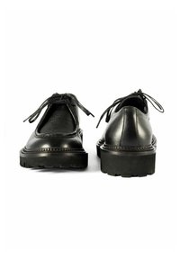 Para czarnych skórzanych butów wiązanych z grubymi, teksturowanymi podeszwami, widziana z przodu i z tyłu na białym tle.