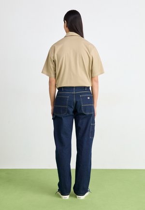 Personne vue de dos portant une chemise beige à manches courtes et un jean bleu foncé avec de grandes poches arrière, debout sur un sol vert clair.