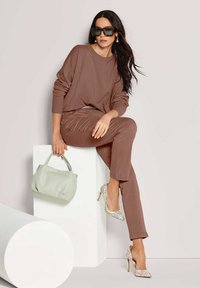 Frau in braunem Pullover und Hose sitzt auf einem weißen Block, hält eine hellgrüne Handtasche, trägt große Sonnenbrillen und gemusterte High Heels.