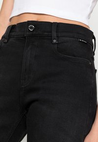 Gros plan sur un jean noir taille haute avec bouton et passants, porté avec un top blanc court dévoilant le ventre.