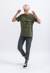 T-shirt en coton vert avec un design graphique de tuyaux et du texte, associé à un pantalon gris fuselé et des baskets noires.