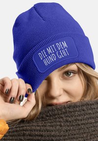 Blue knit beanie with a textured fabric, featuring a rectangular patch that states, “DIE MIT DEM HUND GEHT” in white text.