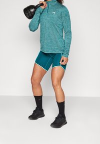 Tealfärgad långärmad träningsblus med dragkedja, matchande tealfärgad shorts med vita detaljer, svarta träningsstrumpor och skor, hållande en svart kettlebell.