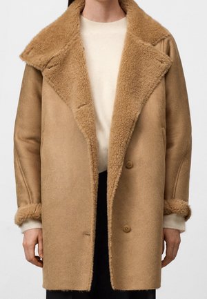 Nainen pukeutunut puolipitkään kamelinnahkaiseen turkistakkiin, jossa on suuret kaulukset, nappikiinnitys ja käännetyt hihat vaaleanneuletakin päällä.