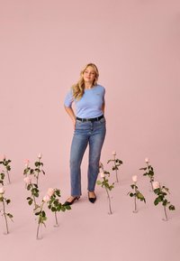 Junge Frau in hellblauem Hemd und Jeans steht zwischen einzelnen rosa Rosen in klaren Vasen auf rosa Boden und Hintergrund.