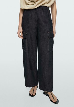 Schwarze, weite Cargohose mit hoher Taille und großen Seitentaschen, kombiniert mit einem beigen Top und schwarzen Zehensandalen auf einfachem Hintergrund.