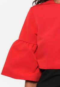 Top rosso corto con maniche a campana, caratterizzato da una texture liscia e dettagli arricciati alle cuciture delle maniche. Parte inferiore nera non visibile.