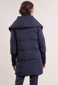 Person med brunt hår i en knut som bär en lång mörkblå vadderad kappa med bred krage och ribbad stickad ärm, står med ryggen mot kameran.