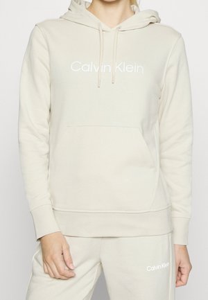 Person trägt beige Calvin Klein Hoodie und passende Jogginghose mit Fronttasche und Kapuze mit Kordelzug.