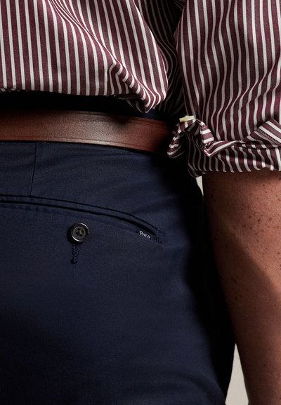 Bordó és fehér csíkos ing, begombolva sötétkék nadrágba, barna bőrpánttal, hátul egy "Polo" feliratú begombolható zsebbel.