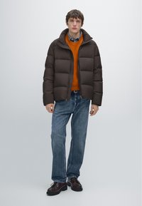 Brązowa pikowana kurtka puchowa z wysokim kołnierzem, noszona na pomarańczowym swetrze, zestawiona z niebieskimi dżinsami i ciemnym obuwiem. Prosty przód i luźny krój.