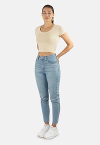Beige Cropped-T-Shirt, hochtaillierte hellblaue Jeans und weiße Sportschuhe. Das Modell steht mit Händen in den Hüften vor einem neutralen Hintergrund.