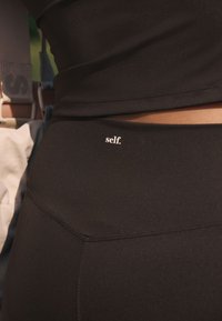 Mustat aktiivitrikoot, joissa on sileä ja joustava materiaali sekä pieni valkoinen logo, jossa lukee "self." vyötärön läheisyydessä. Parannellut saumeiden yksityiskohdat.