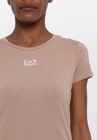 Šviesiai rudas prigludęs marškinėliai, pagaminti iš lygios medžiagos. Turi apvalų kaklą ir trumpas rankoves, ant krūtinės yra nedidelis baltas EA7 logotipas.
