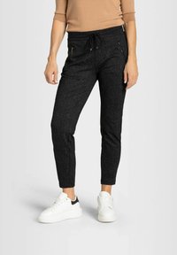 Kvinna klädd i svarta mönstrade slim joggerbyxor med dragkedjefickor, beige topp och vita sneakers som står mot en vit bakgrund.