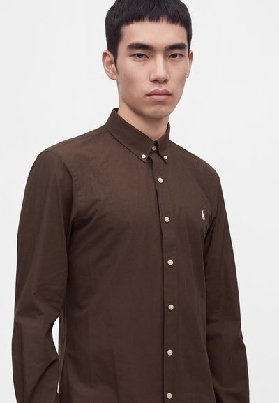 Jeune homme aux cheveux courts et noirs portant une chemise marron foncé boutonnée avec un petit logo blanc sur la poitrine, sur un fond uni.