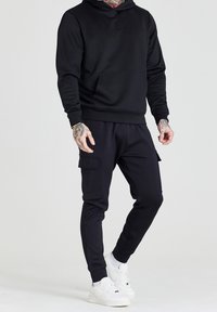 Zwarte hoodie met reliëf tekst, gecombineerd met zwarte cargojoggers met zijzakken, gedragen met witte sneakers.