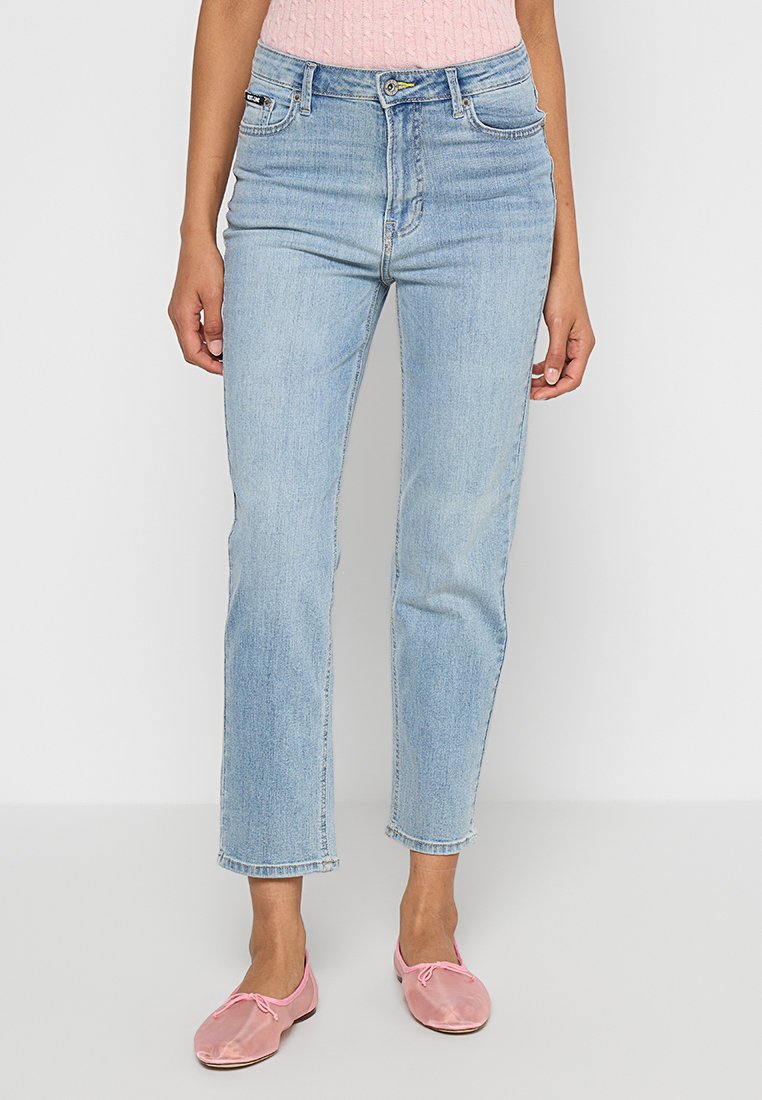 Kobieta w jasnoniebieskich prostych dżinsach, różowej dzianinowej bluzce i satynowych różowych baletkach, stojąca na tle białej ściany.